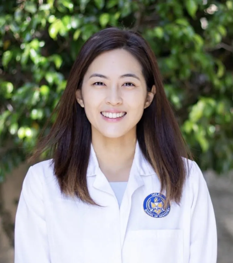 초록 잎이 배경인 옥외에서 흰 가운을 입은 여성 의료진의 headshot 사진.